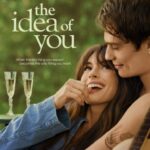 Poster for "The Idea of You." Anne Hathway sits in Nicholas Galitizine's arms. They smile at each other. Glasses of wine are in the background.